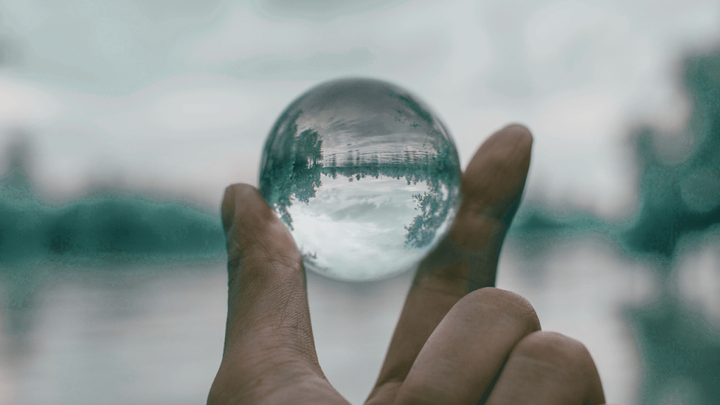 A close-up of a person's hand holding a crystal ball that reflects trees and a cloudy sky.