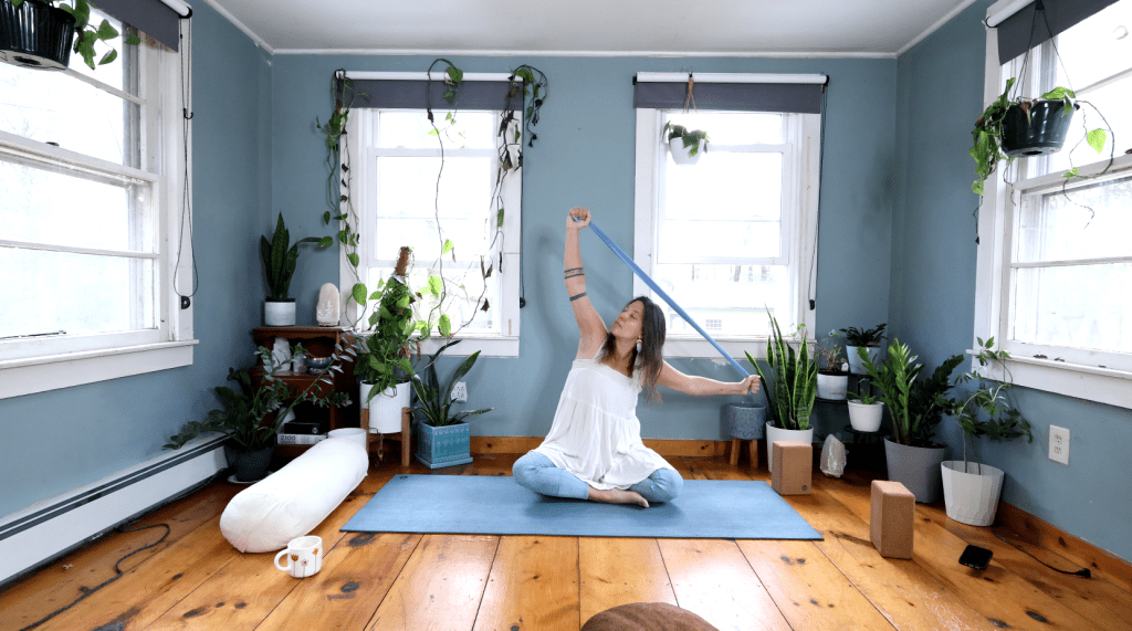 A person practicing yoga in a serene indoor space with plants, wearing a white top and blue jeans, stretching with a yoga strap on a blue mat.