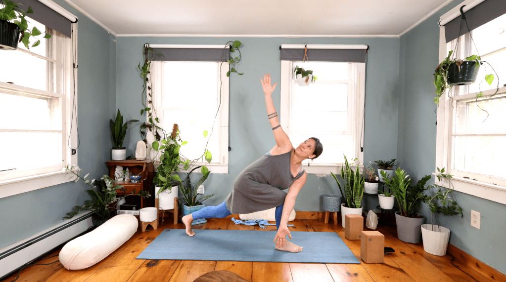 A woman performing a yoga pose in a bright, plant-filled room with wooden floors and large windows.