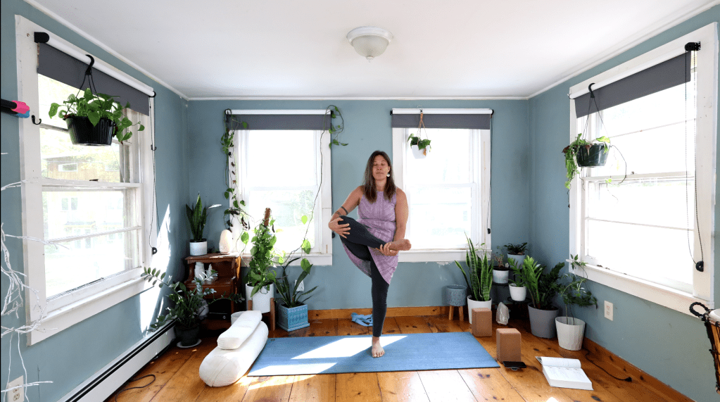 A woman practicing a yoga pose in a bright, plant-filled room with large windows.